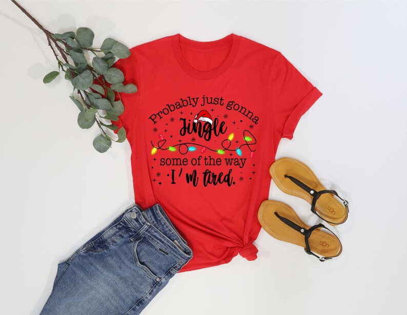 May include: A red t-shirt with the text "Probably just gonna Jingle some of the way, I'm tired." The shirt is paired with blue jeans and tan sandals with black straps. A sprig of green leaves is also present.