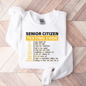 May include: White sweatshirt with a yellow graphic that says "Senior Citizen Texting Code" and lists common texting abbreviations and their meanings.