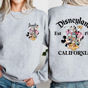 May include: A light gray sweatshirt featuring a colorful graphic of Disney characters. The front has the name "Jessica" above the characters. The back displays "Disneyland California Est 1955" in black script.