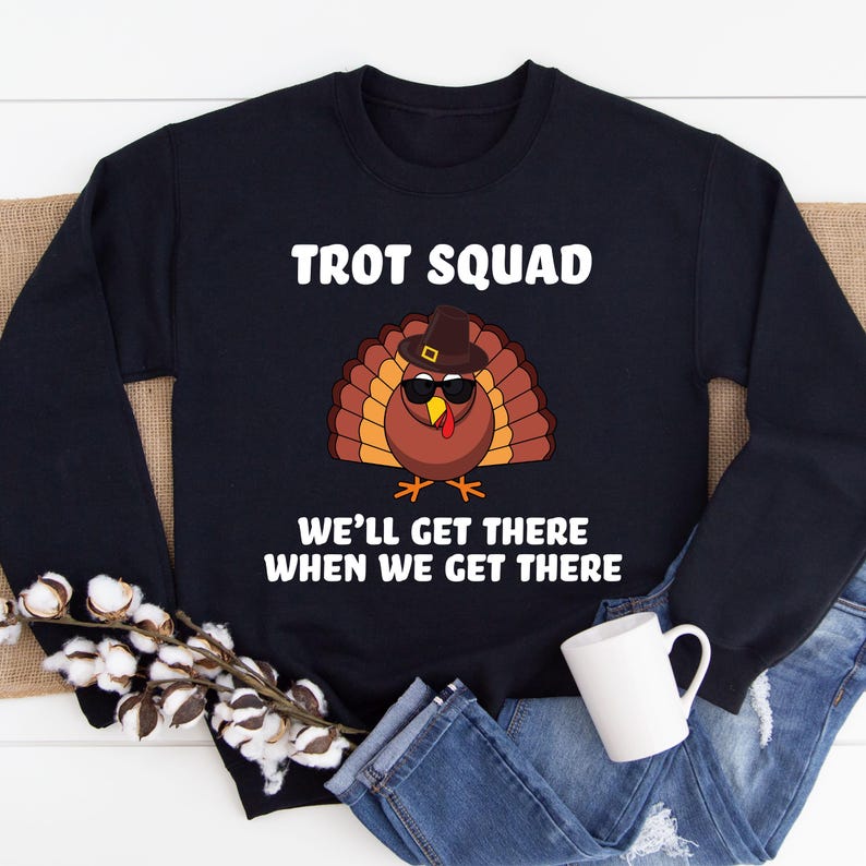May include: A navy blue sweatshirt featuring the words "TROT SQUAD" above a cartoon turkey wearing sunglasses and a hat. The text "WE'LL GET THERE WHEN WE GET THERE" is below the turkey. The sweatshirt is on a white surface.