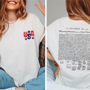 May include: Cream-colored long-sleeve shirt with a USA graphic on the front and the Declaration of Independence printed on the back. The USA graphic features red and blue stars and letters. The back of the shirt displays the text "In Congress, July 4, 1776."