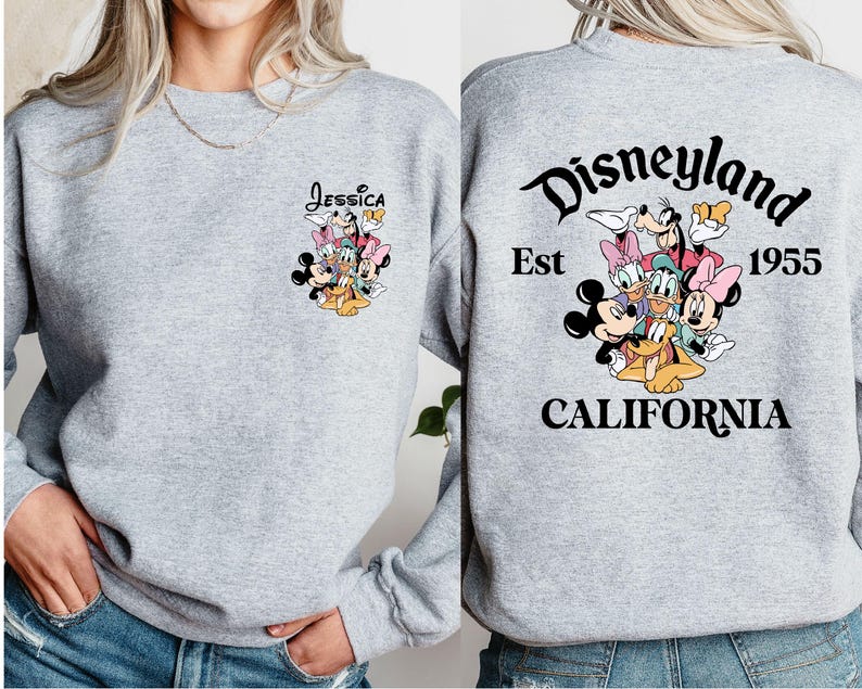 May include: A light gray sweatshirt featuring a colorful graphic of Disney characters on the front and back. The front has the name "Jessica" above the characters. The back displays "Disneyland California Est 1955".