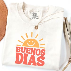 May include: Cream-colored t-shirt with the words "BUENOS DIAS" in red and a smiling sun graphic in orange. A gold necklace with a pendant rests on the shirt. A brown hat and dried palm leaf are in the background.