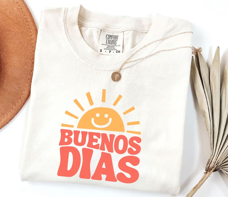 May include: Cream-colored t-shirt with the words "BUENOS DIAS" in red and a smiling sun graphic in orange. A gold necklace with a pendant rests on the shirt. A brown hat and dried palm leaf are in the background.