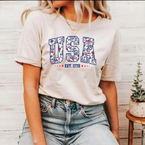 May include: A cream-coloured t-shirt featuring the word "USA" in a floral design, outlined in blue. The text "EST. 1776" is below the word "USA", with red stars on either side. The shirt is a casual style.