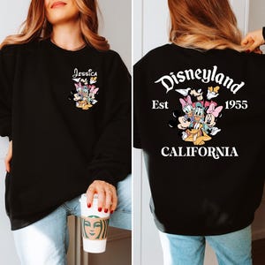 May include: Black sweatshirt featuring a colorful graphic of Disney characters. The front displays the name "Jessica" above the characters. The back of the sweatshirt has the Disneyland logo, the year "1955", and the word "CALIFORNIA".