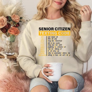 May include: Beige sweatshirt with the text "Senior Citizen Texting Code" in yellow. The text is a list of abbreviations and their meanings, such as BFF, LOL, BTW, TTYL, BYOT, LMOO, WAITT, OMSG, GGPBL, and HOFLACGU.