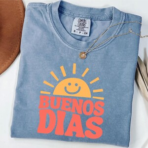 May include: A blue Comfort Colors t-shirt with a smiling sun graphic in orange and the words "BUENOS DIAS" in red. A gold necklace with a small pendant rests on the shirt. The shirt is size S-P-CH.