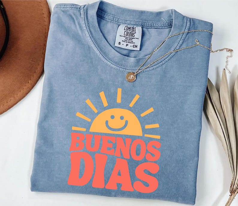 May include: A blue Comfort Colors t-shirt with a smiling sun graphic in orange and the words "BUENOS DIAS" in red. A gold necklace with a small pendant rests on the shirt. The shirt is size S-P-CH.