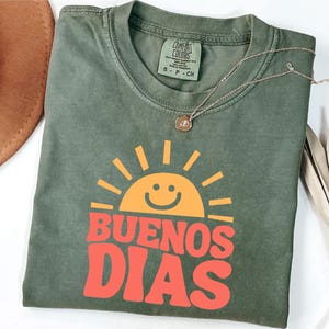May include: Olive green t-shirt with the words "BUENOS DIAS" in orange and a smiling sun graphic. The shirt has a crew neck and short sleeves. A gold necklace with a pendant is draped across the shirt.