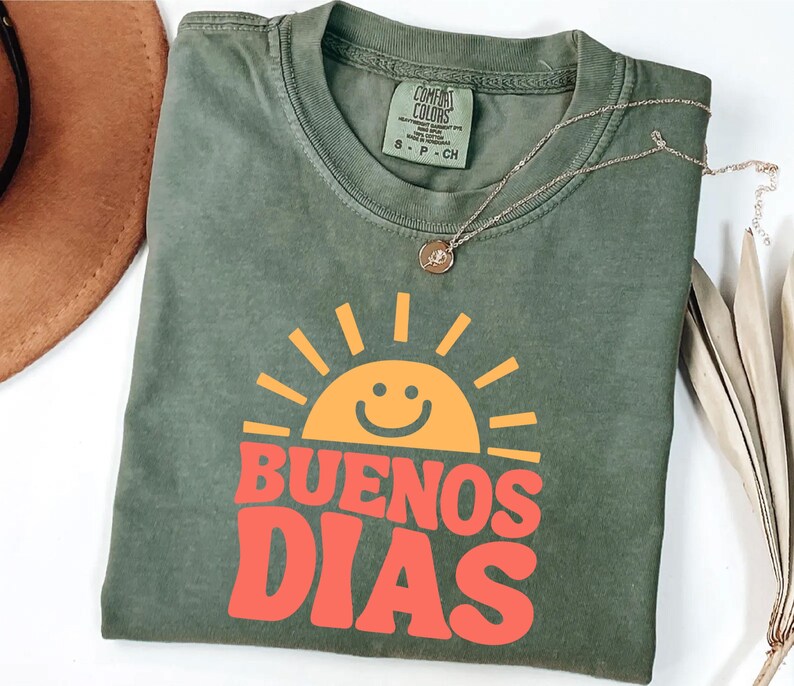May include: Olive green t-shirt with the words "BUENOS DIAS" in orange and a smiling sun graphic. The shirt has a crew neck and short sleeves. A gold necklace with a pendant is draped across the shirt.