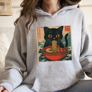 May include: A light gray hoodie featuring a graphic of a black cat eating ramen from a red bowl. The image has Japanese text and a vintage aesthetic. The cat is the focal point, with the word "Ramen" on the left side of the image.