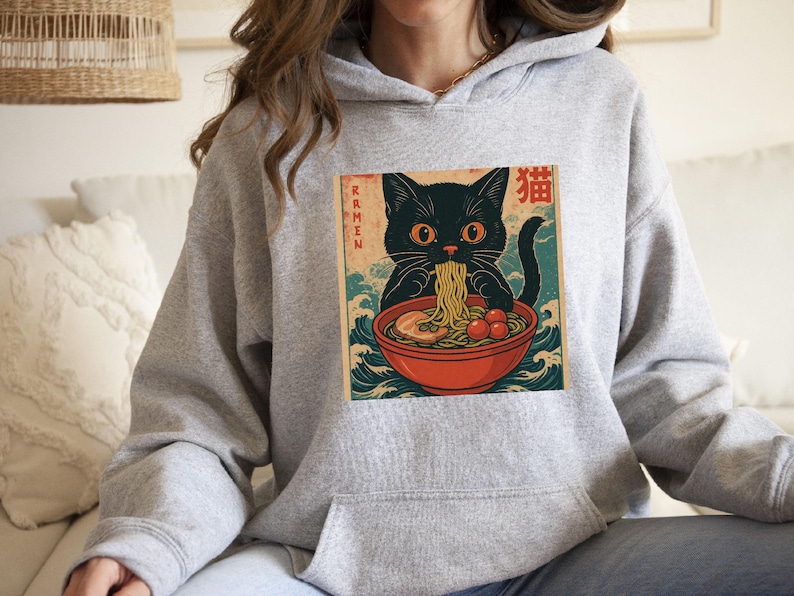 May include: A light gray hoodie featuring a graphic of a black cat eating ramen from a red bowl. The image has Japanese text and a vintage aesthetic. The cat is the focal point, with the word "Ramen" on the left side of the image.