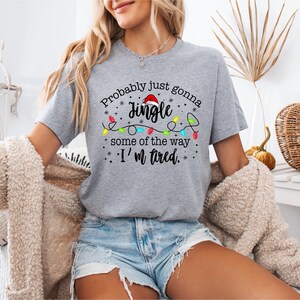 May include: A gray t-shirt with the text "Probably just gonna Jingle some of the way, I'm tired." The design includes a Santa hat and colorful Christmas lights. The shirt is short-sleeved.