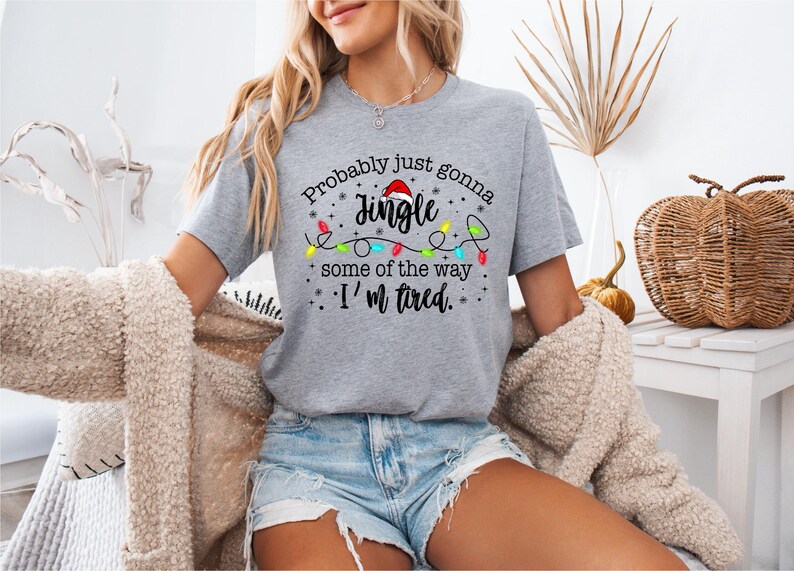 May include: A gray t-shirt with the text "Probably just gonna Jingle some of the way, I'm tired." The design includes a Santa hat and colorful Christmas lights. The shirt is short-sleeved.