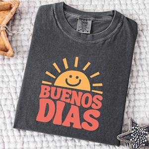 May include: Dark gray t-shirt with a retro design featuring a smiling sun and the words "BUENOS DIAS" in orange. The shirt is folded and lies on a white textured surface. A wooden star-shaped bowl and a beaded star-shaped ornament are in the background.