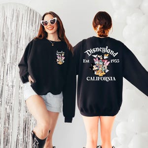 May include: Black sweatshirt featuring a Disneyland graphic on the back with the text "Disneyland Est. 1955 California". The front of the sweatshirt has a smaller graphic with the name "Jessica". The sweatshirt has long sleeves.