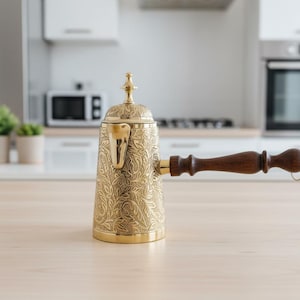 Op de afbeelding: Een goudkleurige, sierlijke koffiepot met een bloemmotief en een donkerhouten handvat. De pot heeft een decoratief deksel en tuit. De achtergrond toont een keukenomgeving met een magnetron en oven.