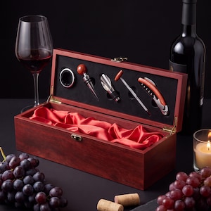May include: A rectangular wooden wine box with a hinged lid, featuring a red satin lining and a foam insert holding a silver-toned wine stopper, pourer, thermometer, and corkscrew. The box is finished in a dark reddish-brown wood grain.