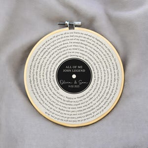 May include: A white embroidery hoop with a black and white record design. The record features the lyrics to the song "All of Me" by John Legend. The text "Olivia & Sam 14.02.2023" is written on the record.