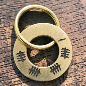 May include: A circular brass keychain charm featuring a brushed metallic finish and a smooth gold-toned split ring. The flat, round charm is engraved with four sets of tally marks and a single horizontal line, creating a distinct numerical pattern.