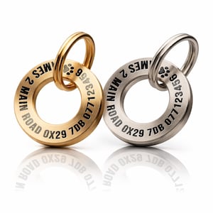 May include: Two metal pet ID tags, one gold-toned, one silver. Each tag has a circular design with a ring. The tags display the text "MES 2 MAIN ROAD OX29 7DB 077123456" and a paw print. Ideal for pet owners.