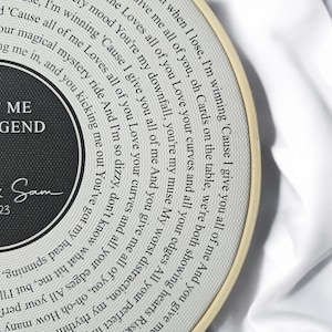 May include: A white and black circular embroidery hoop with a black and white design. The design features a record with the words "You're my downfall, you're my muse My worst distraction, my rhythm" and "ME LEGEND" in black text. The text "Sam 23" is also included in black text.