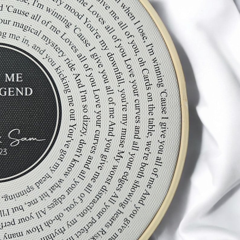 May include: A white and black circular embroidery hoop with a black and white design. The design features a record with the words "You're my downfall, you're my muse My worst distraction, my rhythm" and "ME LEGEND" in black text. The text "Sam 23" is also included in black text.