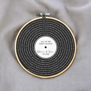 May include: Black and white embroidery hoop with the lyrics to the song "All of Me" by John Legend. The lyrics are arranged in a circular pattern around a white circle with the song title and artist name. The date "14.02.2023" and the names "Olivia & Sam" are written in cursive on the white circle.