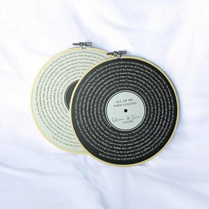 May include: Two black and white vinyl record-style embroidery hoops with the lyrics to the song "All of Me" by John Legend. The text on the hoops reads "All of Me John Legend" and "Olivia & Sam 14.02.2023".