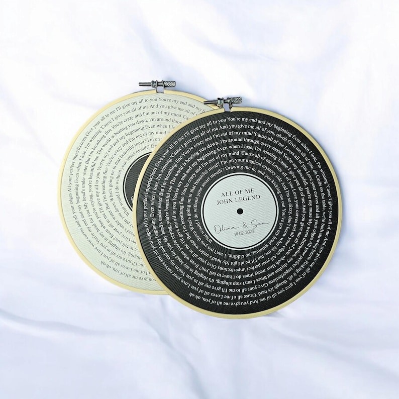 May include: Two black and white vinyl record-style embroidery hoops with the lyrics to the song "All of Me" by John Legend. The text on the hoops reads "All of Me John Legend" and "Olivia & Sam 14.02.2023".