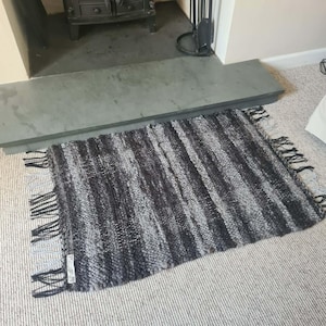 May include: A rectangular, handwoven rug with a mix of grey and black stripes and fringe detailing. The rug is placed on a light-coloured carpet in front of a fireplace, adding texture and warmth to the room.