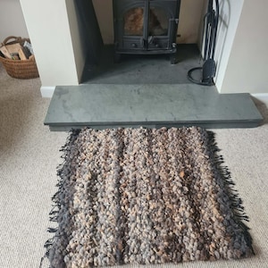 May include: A rectangular rug with a textured surface in shades of brown, grey, and beige. The rug has a shaggy texture and a fringed edge. It is positioned in front of a fireplace.