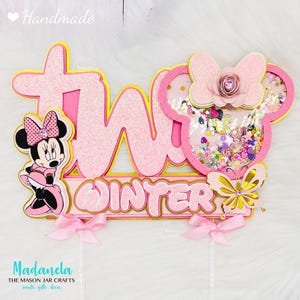 Può includere: Topper per torta in glitter rosa e oro con il numero "due", la parola "Winter", un personaggio di Minnie Mouse, un fiocco rosa con un fiore e una farfalla.