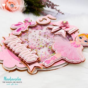 May include: A pink and gold glitter cake topper with a ballerina figurine, a pink bow, and the name "Victoria" written in pink and white letters. The topper also includes the number "5" in pink and gold.
