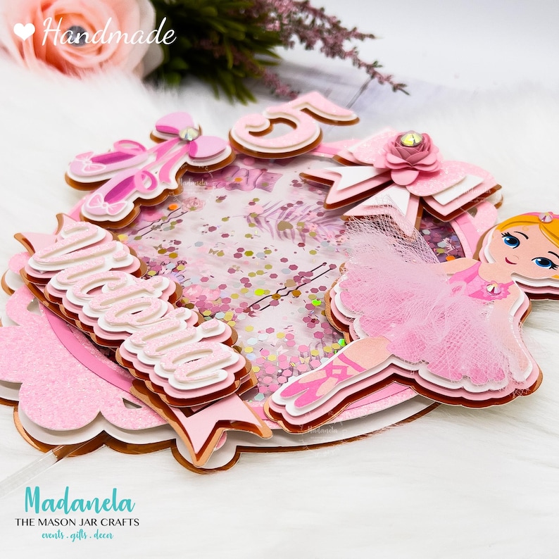 May include: A pink and gold glitter cake topper with a ballerina figurine, a pink bow, and the name "Victoria" written in pink and white letters. The topper also includes the number "5" in pink and gold.