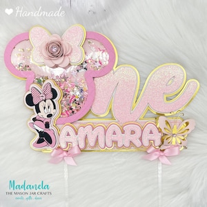 Könnte beinhalten: Handgefertigter rosa-goldener Glitzer-Kuchenaufsatz mit dem Wort „One“ und dem Namen „Amara“. Mit Minnie Mouse-Design, Schmetterling und rosa Schleife. Der Aufsatz hat ein Shaker-Element mit Konfetti und einer Papierblume.
