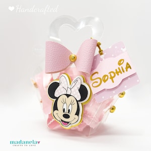Può includere: Un sacchetto regalo trasparente con un fiocco rosa e una grafica di Minnie Mouse. La borsa ha un manico a forma di cuore e un'etichetta con "Sophia" in lettere dorate. La borsa è piena di oggetti rosa.