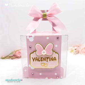 Puede incluir: Una caja de regalo rosa y dorada con tapa transparente. La caja tiene un lazo rosa en la parte superior y un lazo rosa con purpurina en la parte delantera. La caja dice "Valentina" en letras doradas.