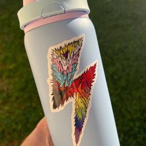 May include: A light blue water bottle with a two-toned light blue and pink lid. A sticker featuring a jagged, lightning bolt-shaped design with a multi-coloured, spiky, and textured pattern in shades of yellow, pink, blue, red, orange, and purple.