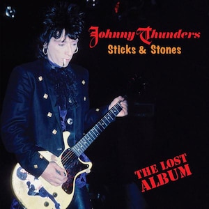 Puede incluir: Una fotografía en blanco y negro de un hombre tocando una guitarra. Lleva una chaqueta negra y una camisa blanca. La guitarra es de color marrón claro con una placa protectora negra. El texto "Johnny Thunders Sticks & Stones The Lost Album" está en letras rojas sobre un fondo negro.