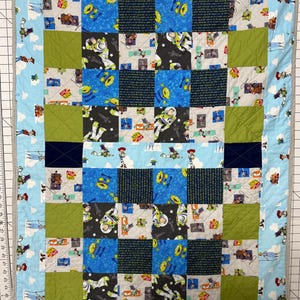Op de afbeelding: Een handgemaakte patchworkquilt met een blauwe rand met stripfiguren en wolken. Het midden van de quilt heeft een dambordpatroon met vierkanten van blauwe, groene en zwarte stof, met verschillende karakterprints. De quilt is ongeveer 100 cm bij 127 cm groot.