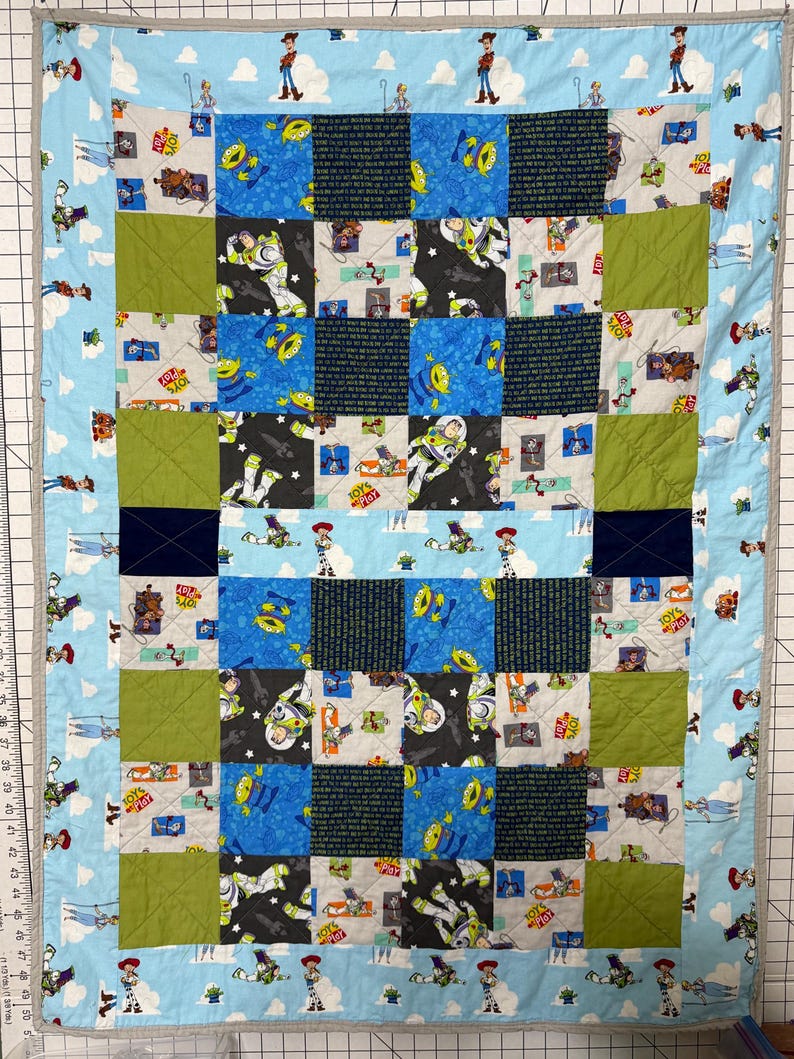 Op de afbeelding: Een handgemaakte patchworkquilt met een blauwe rand met stripfiguren en wolken. Het midden van de quilt heeft een dambordpatroon met vierkanten van blauwe, groene en zwarte stof, met verschillende karakterprints. De quilt is ongeveer 100 cm bij 127 cm groot.