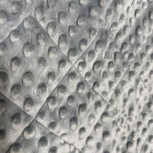Op de afbeelding: Close-up van een lichtgrijze, pluche deken met een gewatteerd ruitpatroon. De stof heeft een zacht, getextureerd oppervlak met verhoogde, cirkelvormige stippen. De deken lijkt gemaakt te zijn van een zacht, donzig materiaal.
