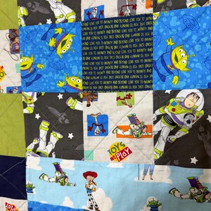 Op de afbeelding: Een patchworkquilt met verschillende personages uit een populaire animatiefilm. De quilt toont afbeeldingen van Buzz Lightyear, aliens en andere personages op een blauwe, groene en grijze achtergrond. De zin "Toys at Play" is ook zichtbaar.