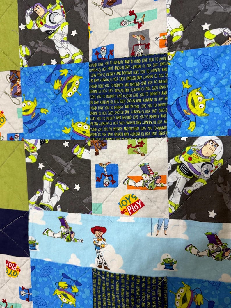 Op de afbeelding: Een patchworkquilt met verschillende personages uit een populaire animatiefilm. De quilt toont afbeeldingen van Buzz Lightyear, aliens en andere personages op een blauwe, groene en grijze achtergrond. De zin "Toys at Play" is ook zichtbaar.