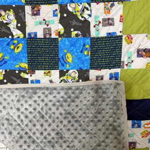 Op de afbeelding: Een patchworkquilt met vierkanten met Toy Story-personages, waaronder Buzz Lightyear en aliens. De quilt heeft een grijze, getextureerde achterkant. De vierkanten zijn in verschillende kleuren, waaronder blauw, groen en zwart. De woorden "TOYS" en "PLAY" zijn zichtbaar.