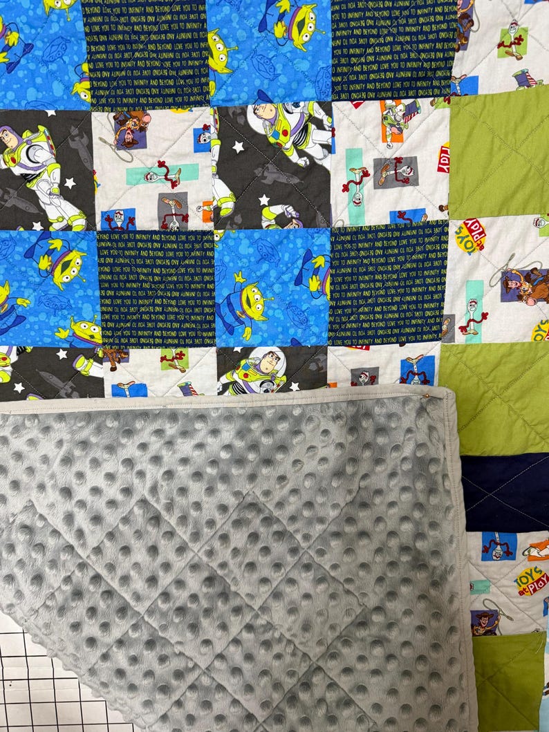 Op de afbeelding: Een patchworkquilt met vierkanten met Toy Story-personages, waaronder Buzz Lightyear en aliens. De quilt heeft een grijze, getextureerde achterkant. De vierkanten zijn in verschillende kleuren, waaronder blauw, groen en zwart. De woorden "TOYS" en "PLAY" zijn zichtbaar.