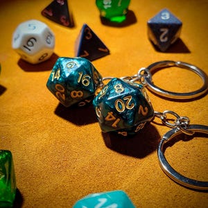 May include: A set of two green and gold dice with black numbers on them. The dice are attached to a silver key chain with two silver rings. The dice are on a brown surface.