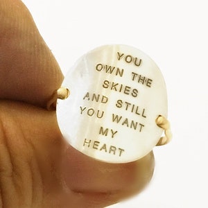 Pu&ograve; includere: Un anello d'oro con una pietra ovale bianca. La pietra &egrave; incisa con le parole "You own the skies and still you want my heart".